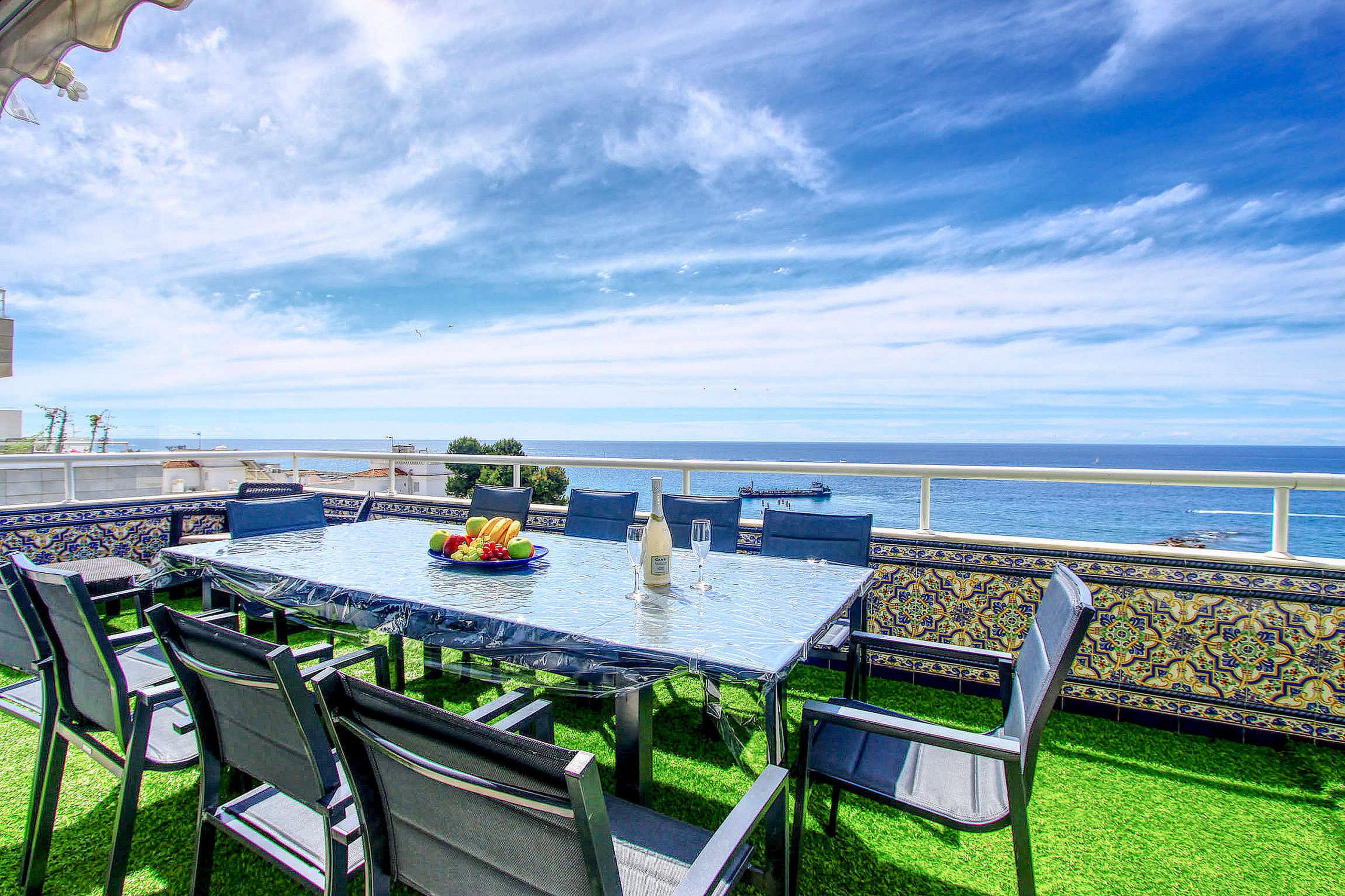 Un espace extérieur aménagé pour les repas, avec vue sur la mer, une table dressée avec une bouteille et des fruits, et des chaises dotées de coussins à motifs.