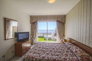 Een hotelkamer met een bed, een televisie op een houten dressoir, een balkon met uitzicht op zee en een kamer met een patroon.