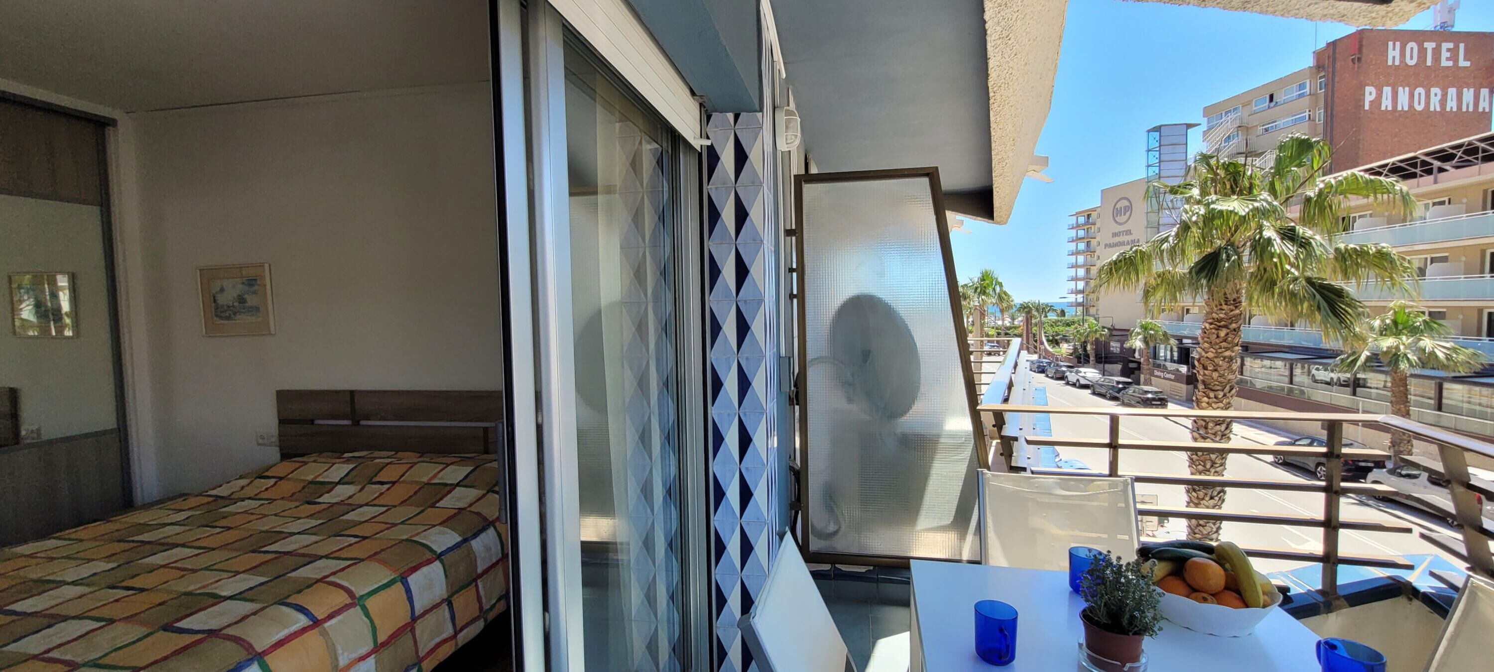 A hotel room with a bed, a balcony with a table, and a view of the street and palm trees.