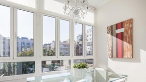 A modern dining area with a glass table and white chairs, large windows with a city view, and a chandelier.