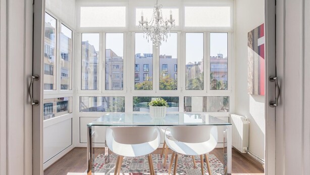 A bright dining area with a glass table, two chairs, a chandelier, and a view of buildings through large windows.