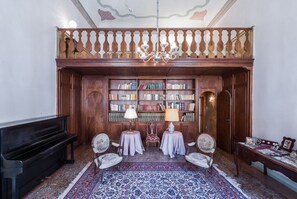 A library with a piano, bookshelves, two tables with lamps, and ornate chairs.