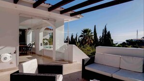 Ein sonnendurchfluteter Sitzbereich im Freien mit einer Couch und Sesseln, eine Glastür, die auf eine Terrasse führt, und Blick auf das Meer und Palmen.