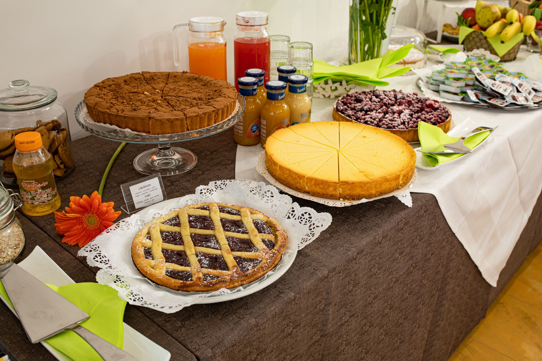 A buffet table with various desserts, including a chocolate cake, a cheesecake, and a pie, along with condiments and beverages.