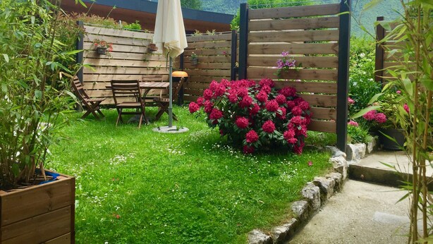 Un jardin avec une table et des chaises en bois, un parasol blanc et un parterre de fleurs.