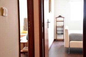 Ett hotellrum med en dörr av trä, en säng med ett rutigt överkast, en vit stol och ett litet sidobord av trä.