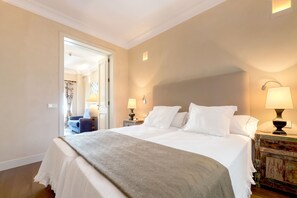 Una camera d'albergo con un letto grande, due lampade sui comodini, un comodino in legno e una porta che dà su un'altra stanza.