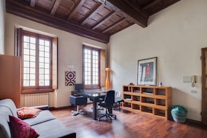 A room with a wooden ceiling, two windows with wooden frames, a desk with a chair, a bookshelf, and a radiator.