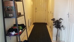 A hallway with a black mat leading to a white door, a metal shelf with bags and a box, and a potted plant on the right. - BEAUTIFUL  BRIGHT MODERN GROUND FLOOR APARTMENT,PARKING, WIFI. (Buckshaw Village)