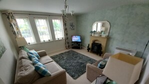 A living room with a beige sofa, a small table, a television, and a fireplace. - BEAUTIFUL  BRIGHT MODERN GROUND FLOOR APARTMENT,PARKING, WIFI. (Buckshaw Village)