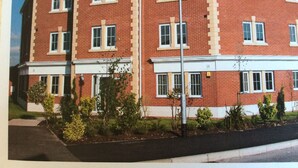A multi-story brick building with a flat roof, several windows, and a small garden area with plants and a street lamp. - BEAUTIFUL  BRIGHT MODERN GROUND FLOOR APARTMENT,PARKING, WIFI. (Buckshaw Village)