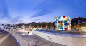 A waterfront promenade with a colorful glass cube structure, a white building with perforated metal detailing, and a distant view of a hilltop building.