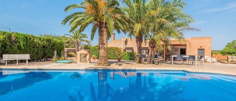 A pool with palm trees and outdoor seating, surrounded by a building with a tiled roof and a stone wall.