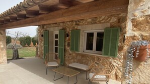 A stone building with a covered patio, two wicker chairs, and a window with green shutters. - Son Coves Rural Cottage (Santanyi)