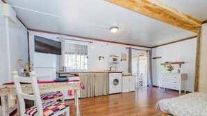 Ein kompakter, gut beleuchteter Raum mit Holzboden, einem Bett, einem Esstisch mit Stühlen, einer Waschmaschine und einer kleinen Küchenzeile mit Fenster.