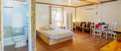 A room with a wooden floor, a bed with a patterned bedspread, a desk with chairs, and a window with curtains.