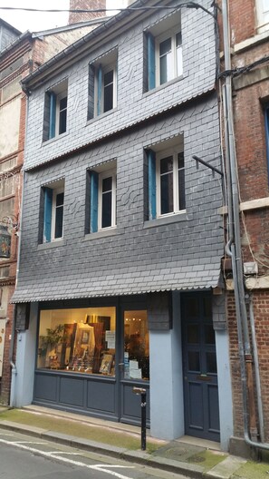 A two-story building with a grey facade, multiple windows with blue shutters, and a ground-floor shop with large glass windows.