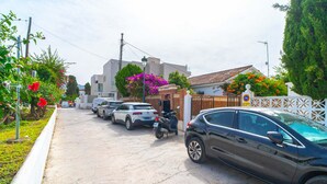 Une rue résidentielle bordée de voitures garées et d’un scooter, des maisons avec des balcons apparents et une clôture blanche.