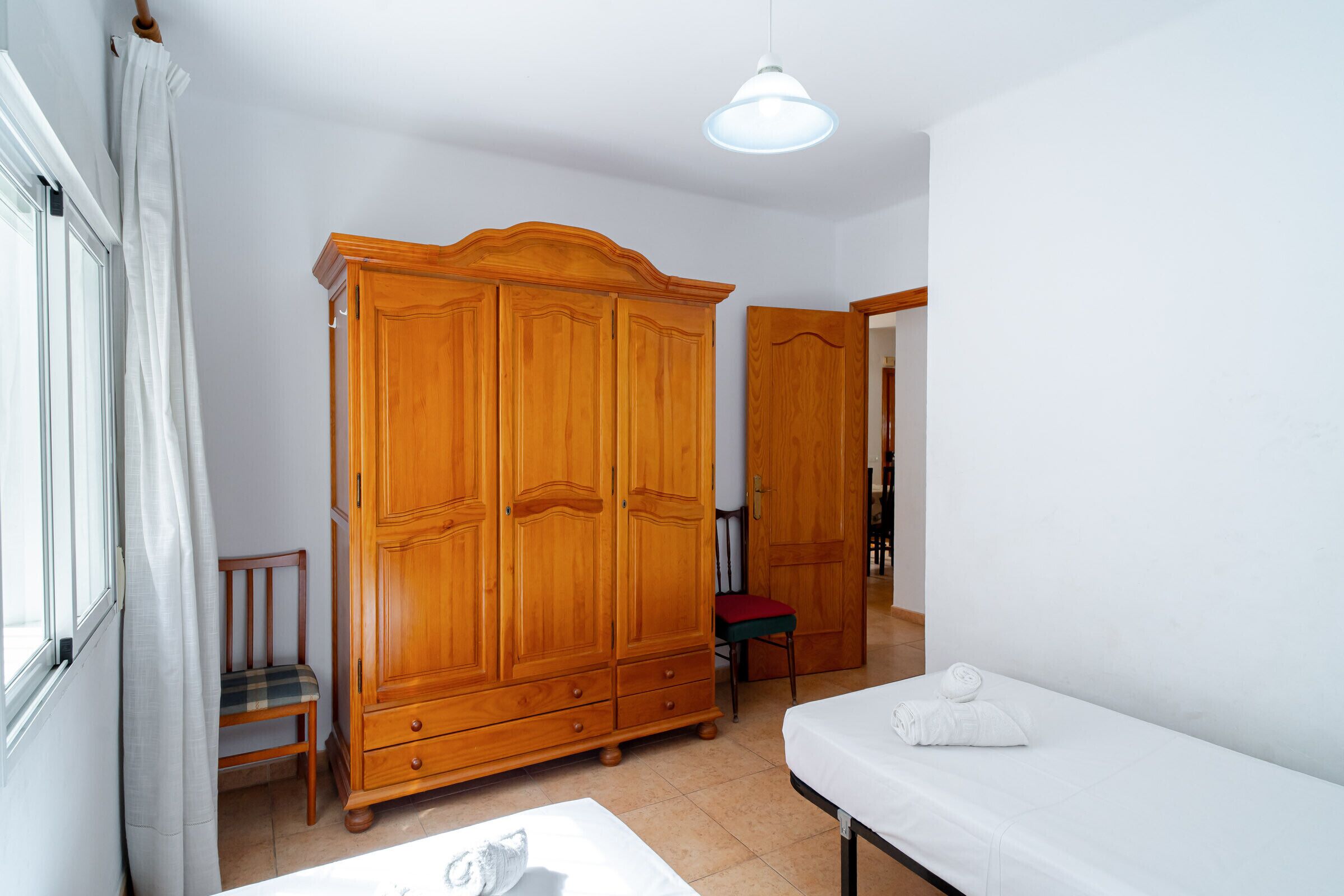 A room with a wooden wardrobe, a chair, a bed with a white mattress, and a hanging light fixture.