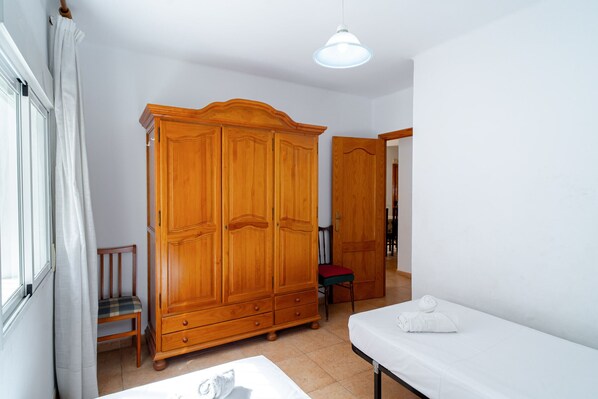 Une pièce comprenant une armoire en bois, une chaise, un lit avec un matelas blanc et un luminaire suspendu.
