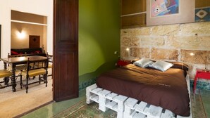 A room with a bed, a wooden table with chairs, a green wall, and a stone wall with a painting.