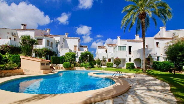 A swimming pool with a stone border, surrounded by white buildings with red-tiled roofs, and a palm tree.