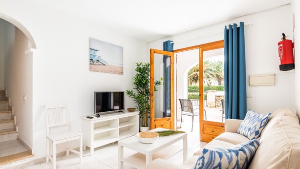 Een woonkamer met een bank, een salontafel, een televisie en een plant. Een deur leidt naar een buitenruimte met uitzicht op palmbomen.