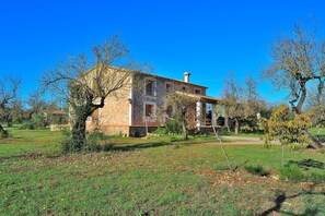 A rustic stone house with a tiled roof, surrounded by a grassy field and trees.
