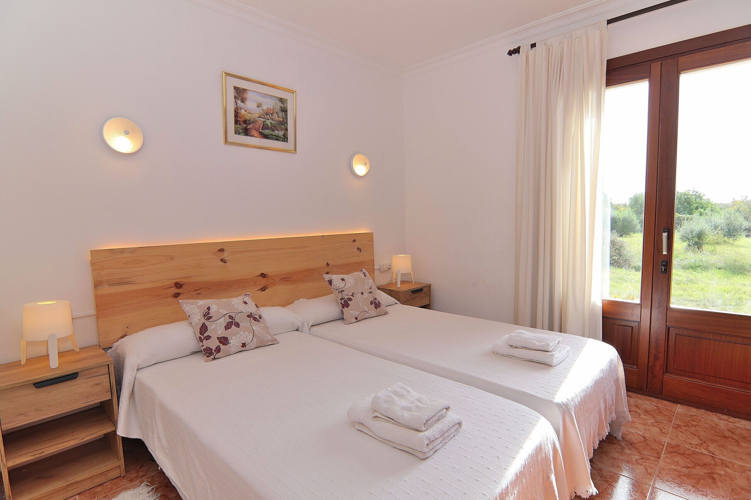 A hotel room with two beds, a wooden headboard, bedside lamps, a framed picture on the wall, and a view through a door.