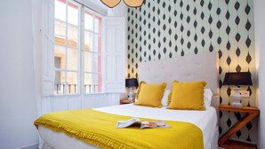 A neatly made bed with a yellow bedspread and pillows, a window with white shutters, and a patterned wallpaper. - Genteel Home Galera Terrace II (Seville)