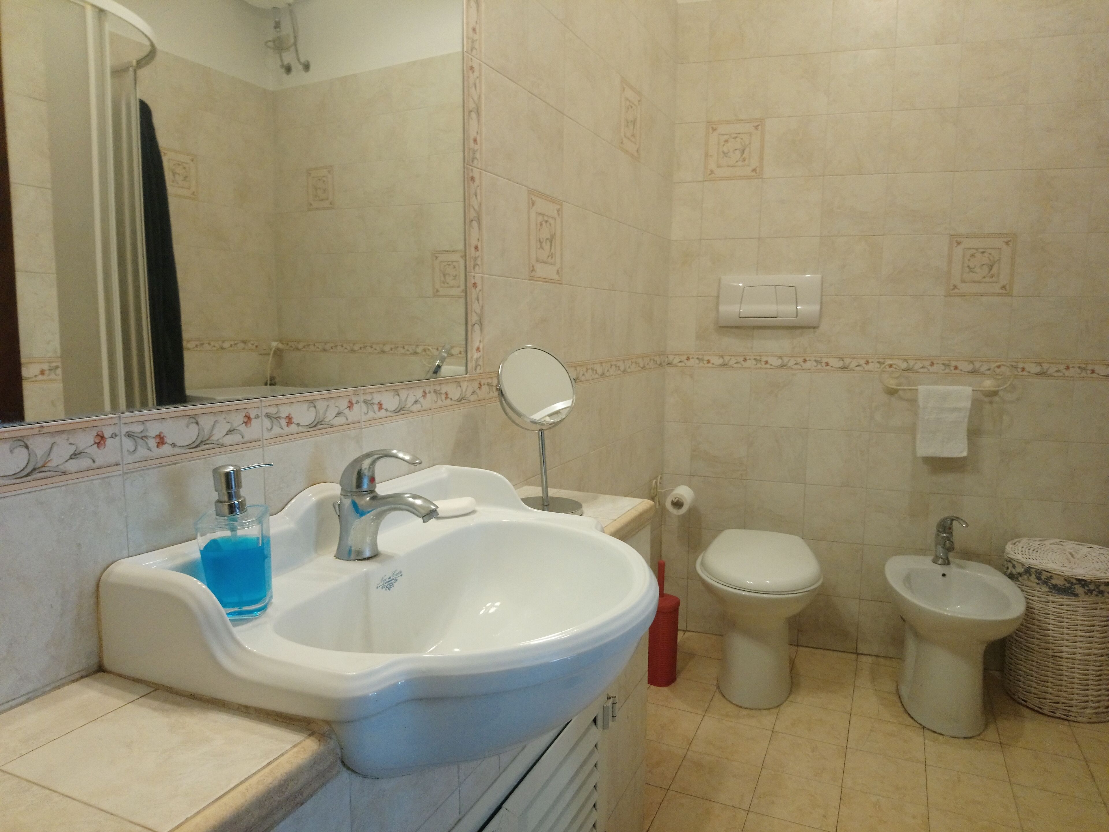 A bathroom with a sink, mirror, toilet, bidet, and a trash bin.