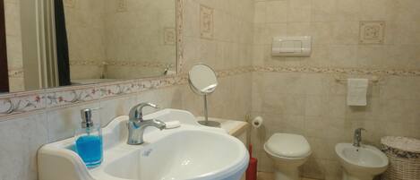A bathroom with a sink, mirror, toilet, bidet, and a trash bin.