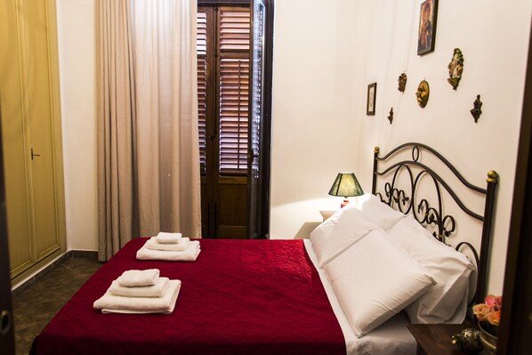 A hotel room with a red bedspread, white pillows, and a metal headboard. There are towels on the bed and a lamp on the nightstand.