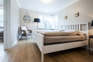 A bedroom with a white bed, a wicker chair, a round mirror, and a window with a view of trees.