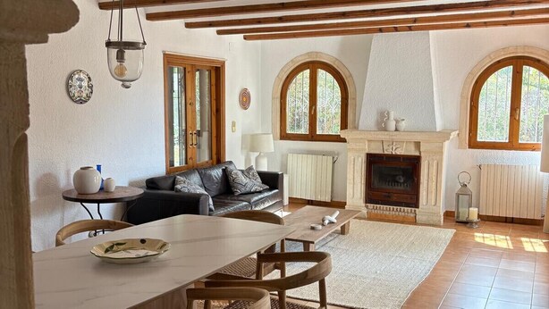 Ein gemütliches Wohnzimmer mit Kamin, Holzbalken, einem Essbereich mit Tisch und Stühlen sowie einem großen Fenster mit Rundbogen.
