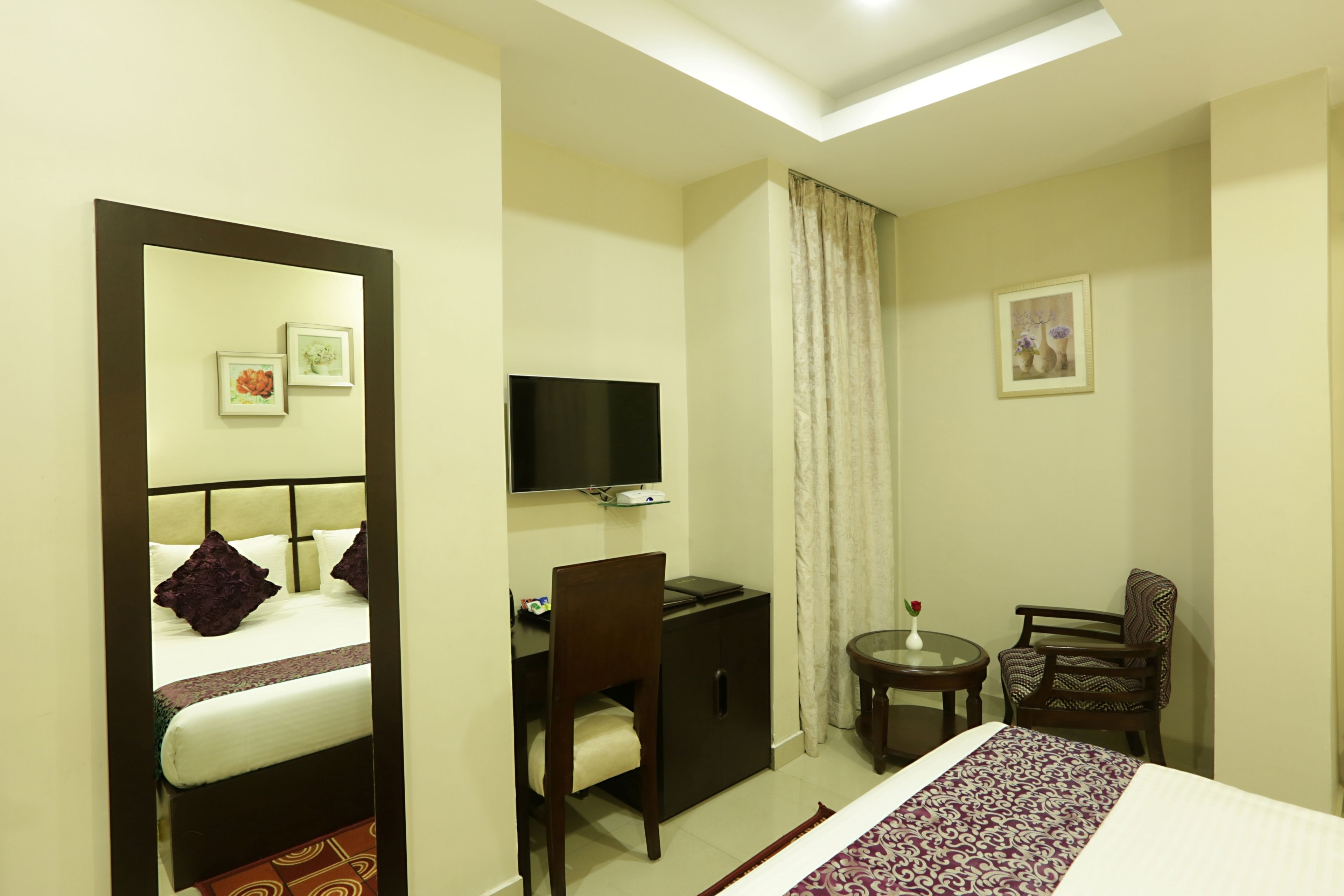 A hotel room with a bed, a desk, a chair, a TV, and a small table with a vase.