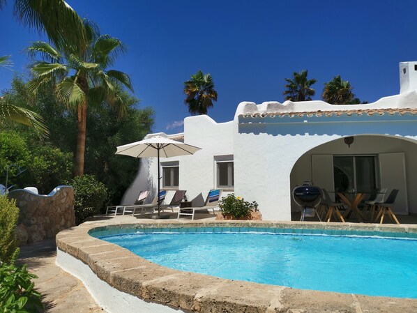 A pool area with a white building, lounge chairs, and a covered seating area.