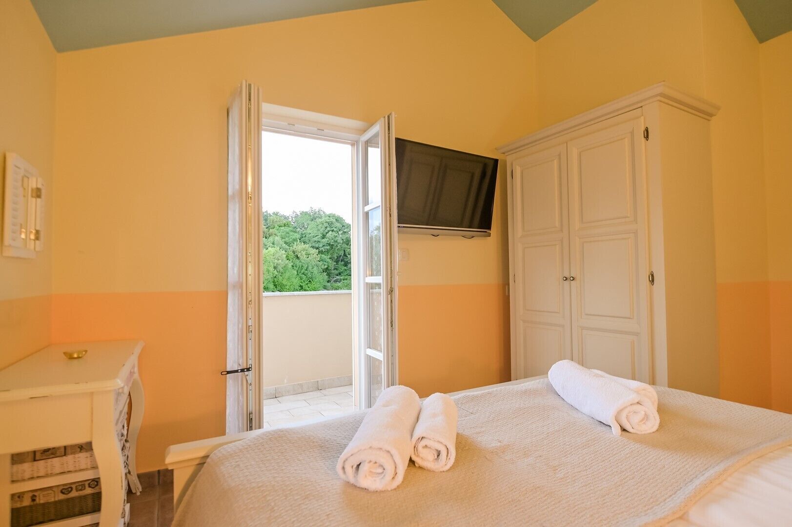 A hotel room with a bed, a TV mounted on the wall, a wardrobe, and an open door leading to a balcony with a view of greenery.