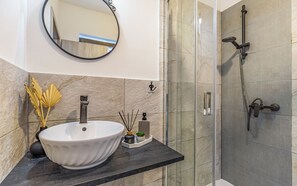 Un baño con un lavamanos blanco tipo lavabo, un espejo redondo y una zona de ducha con mampara de vidrio.