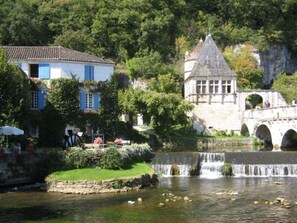 Un puente de piedra histórico sobre un río, una pequeña cascada y edificios con contraventanas azules.