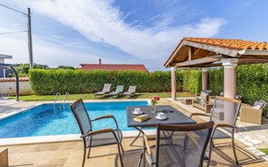A swimming pool with lounge chairs and a table, surrounded by greenery and a building with a red roof in the background. - Villa Sandra 4+2 with pool, garden & terrace with barbecue & free Wi-Fi (Ližnjan)