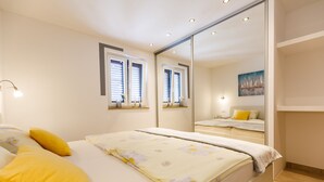 A bedroom with a bed, two windows with blinds, a bedside table, a mirror, and a painting on the wall.