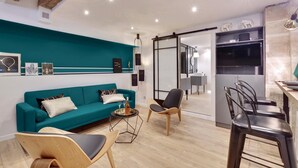 Ein modernes Wohnzimmer mit einem petrolfarbenen Sofa, Holzstühlen, einem kleinen Couchtisch und einer Küchenzeile mit Fernseher und Schränken.