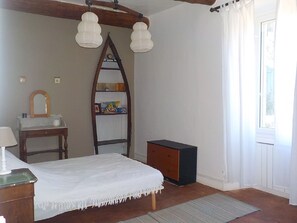 Ein Schlafzimmer mit Bett, Nachttisch, Schminktisch, Spiegel, einer Bücherablage und einem Fenster mit Vorhängen.