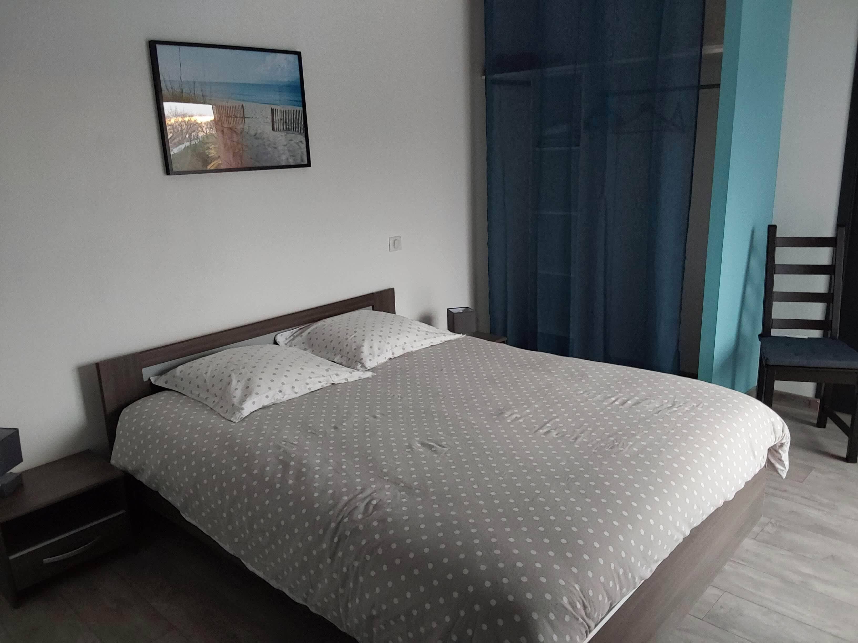 A neatly made bed with a polka dot bedspread and pillows, a bedside table with a lamp, a chair, and a framed picture on the wall.