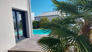 A modern house with a glass door leading to a swimming pool surrounded by lush greenery.