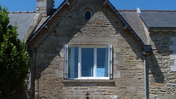 Il s’agit d’un bâtiment en pierre doté d’un toit à deux versants, d’une cheminée et d’une petite fenêtre circulaire. On y voit une porte surmontée d’une fenêtre, et une chaise est visible à l’extérieur.