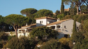 Una villa de estilo mediterráneo con tejado de teja, rodeada de exuberante vegetación y altos árboles.
