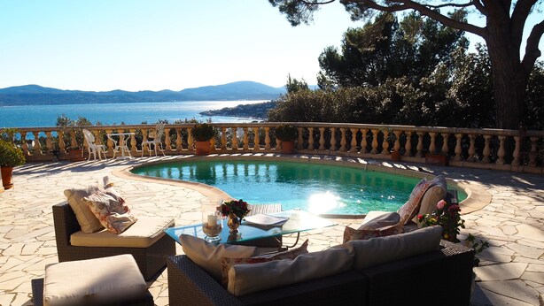 Un área junto a la piscina con mobiliario de mimbre, una mesa de vidrio y vistas al mar y a las montañas.