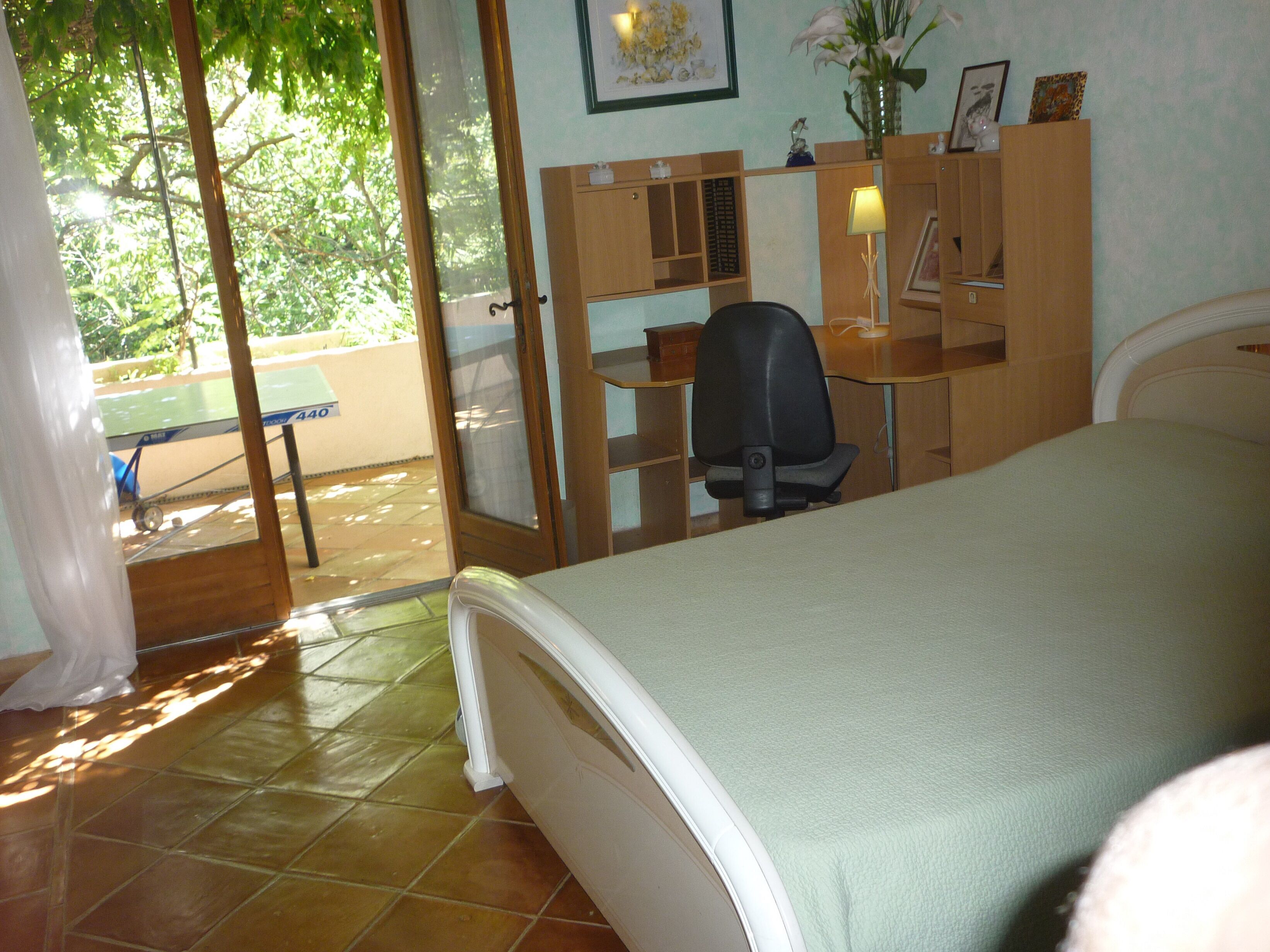 Ein Schlafzimmer mit einem Bett, einem Schreibtisch und einem Stuhl. Es gibt ein Fenster mit Blick auf Bäume und eine Tür zu einem Außenbereich.
